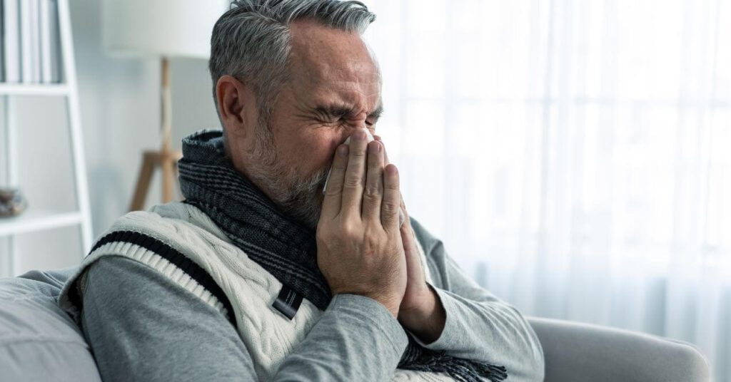 Older man sitting indoors, covering his nose and mouth with his hands, appearing to sneeze or react to cold or allergies.