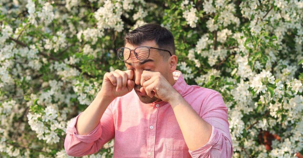 Man standing outside in front of blooming flowers, rubbing his eyes, likely experiencing discomfort due to allergies or irritation.