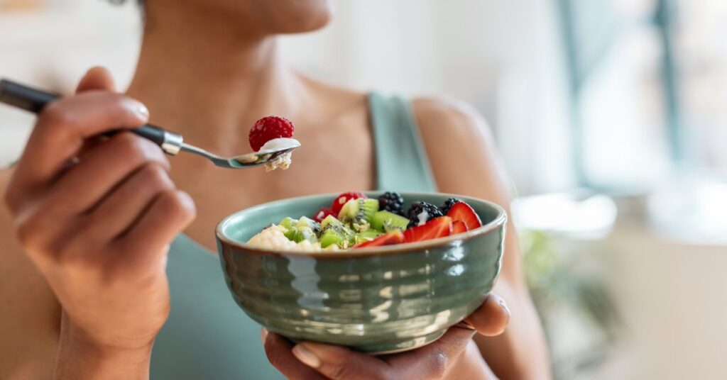 Woman holding a spoon and eating a healthy fruit bowl with strawberries, kiwi, blackberries, and raspberries.