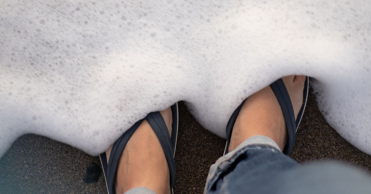 Close-up of feet wearing sandals near the shoreline, surrounded by foam, symbolizing summer footwear.