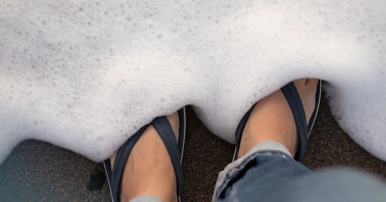 Close-up of feet wearing sandals near the shoreline, surrounded by foam, symbolizing summer footwear.