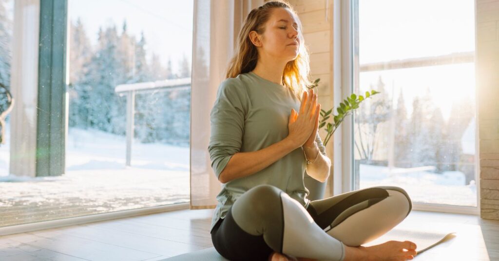 Woman practicing meditation in a peaceful room with large windows and snowy scenery outside, sitting cross-legged with hands in prayer position.