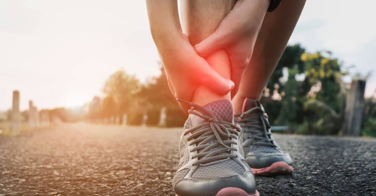 Close-up of a person outdoors, holding their ankle in pain, possibly due to a twisted or sprained ankle while wearing sneakers.