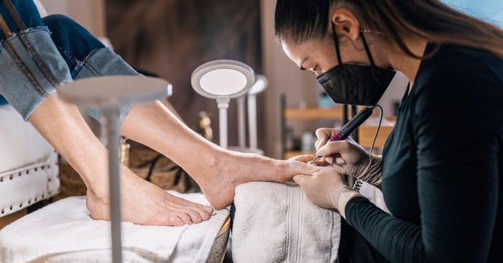 Nail technician wearing a mask and gloves performing a pedicure with a nail drill on a client's foot.