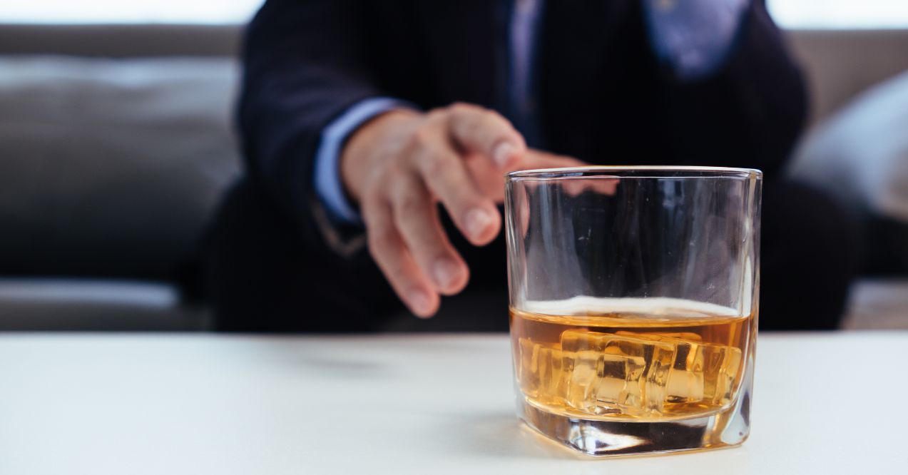 Person reaching for a glass of whiskey on a table, implying alcohol consumption.