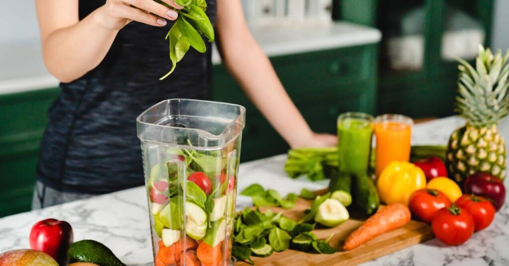 Woman placing fresh leafy greens into a blender surrounded by fruits and vegetables on a kitchen counter.