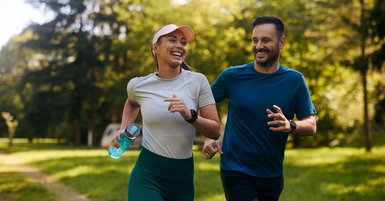 A smiling couple running outdoors on a sunny day in a park, wearing athletic clothes and holding water bottles.