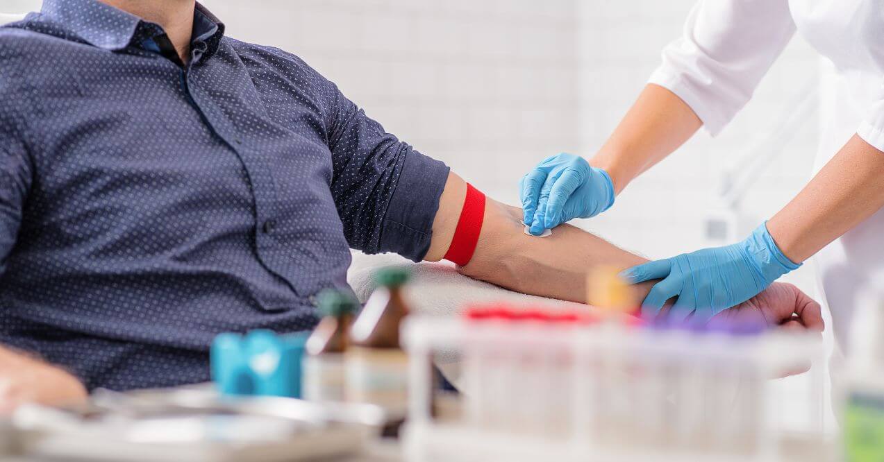 A person getting blood drawn by a healthcare professional wearing gloves, preparing the arm with a red tourniquet.