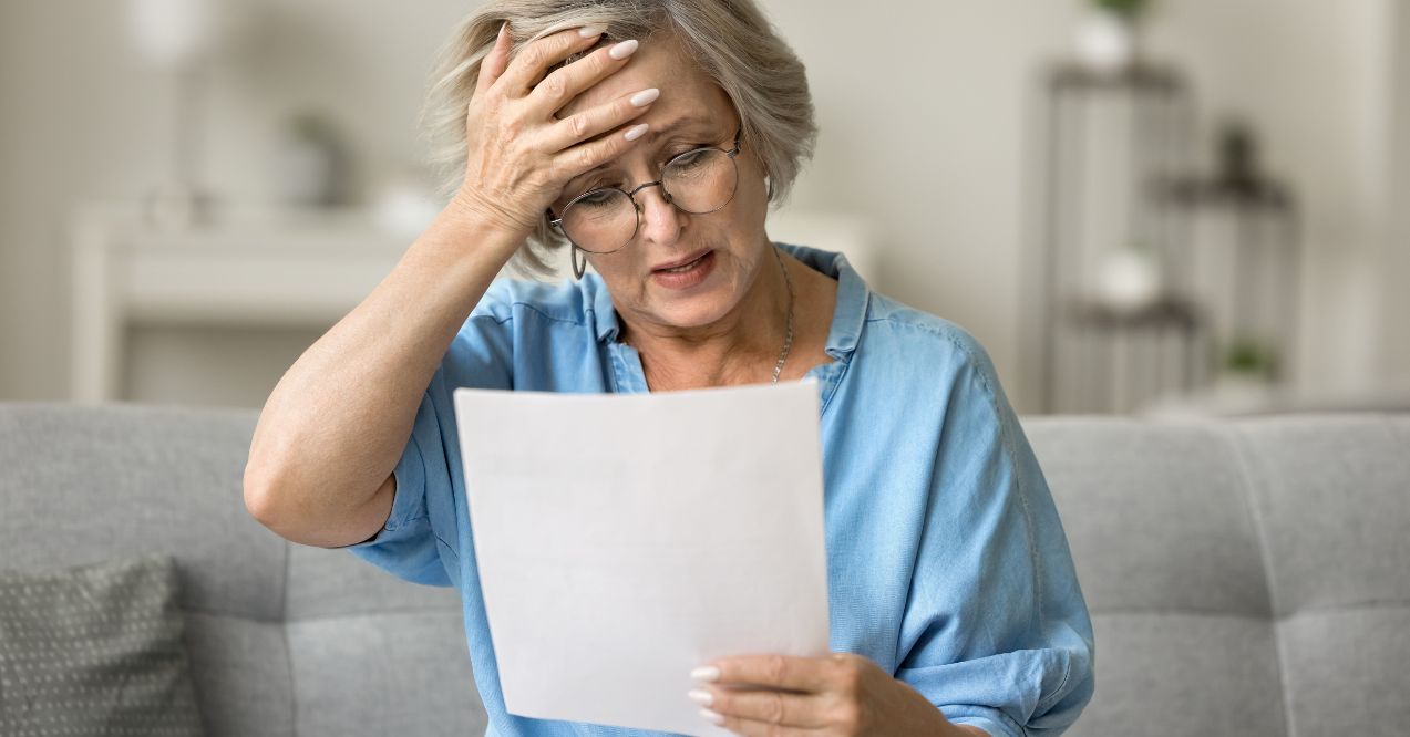 An elderly woman looking stressed and concerned while reading a document in her living room.