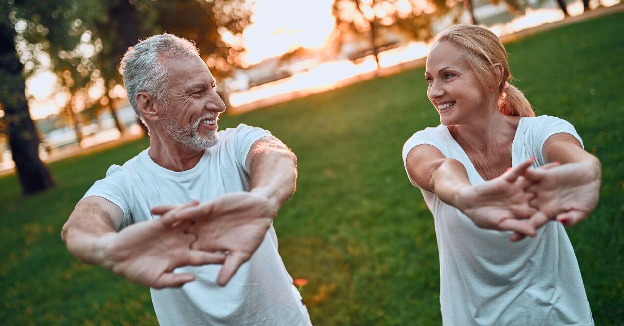 Older couple stretching outdoors and smiling in a park.