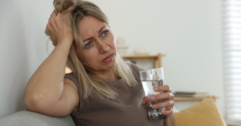 Middle-aged woman holding a glass of water, looking distressed and fatigued.