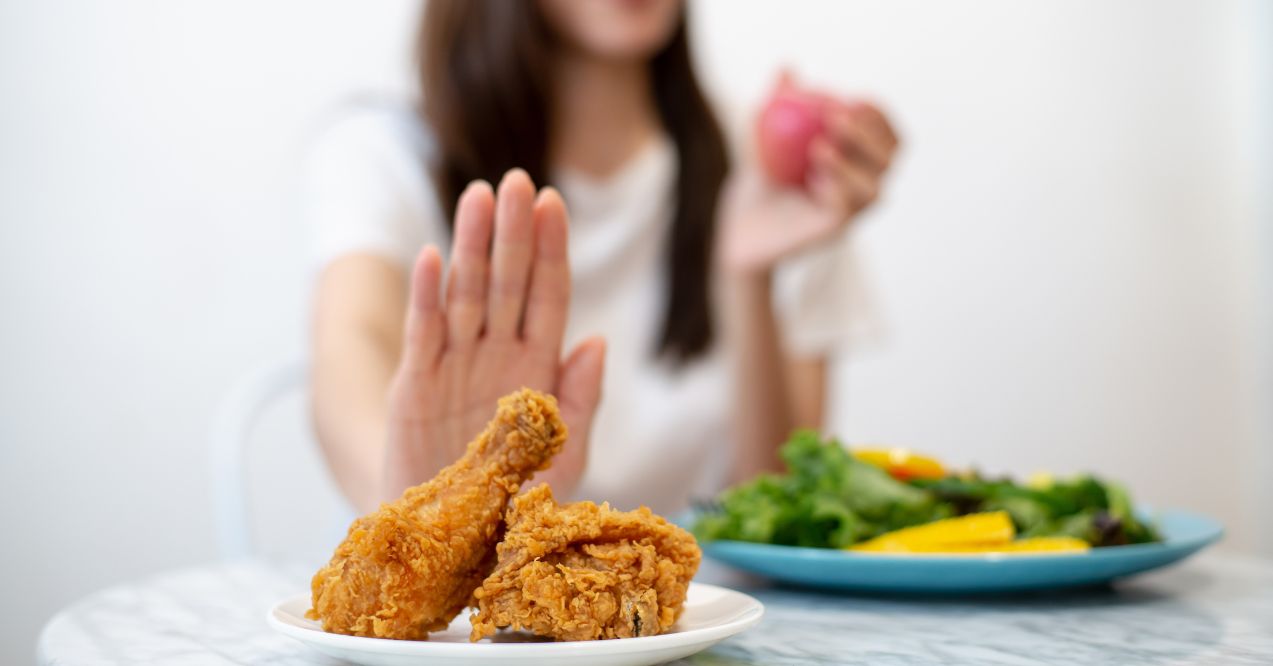 A woman rejecting fried chicken with her hand while sitting at a table, choosing a healthy salad instead.