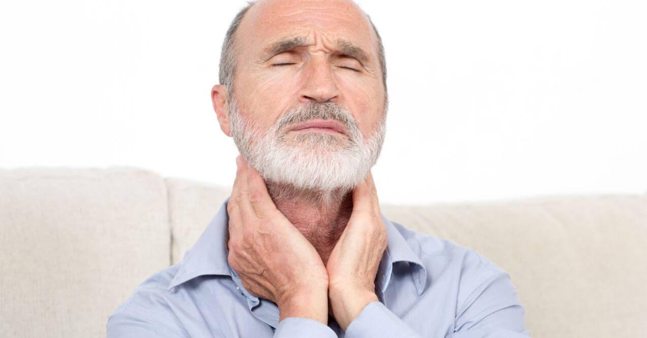 Elderly man sitting on a sofa with his hands on his neck, showing discomfort, related to swelling of preauricular lymph nodes.