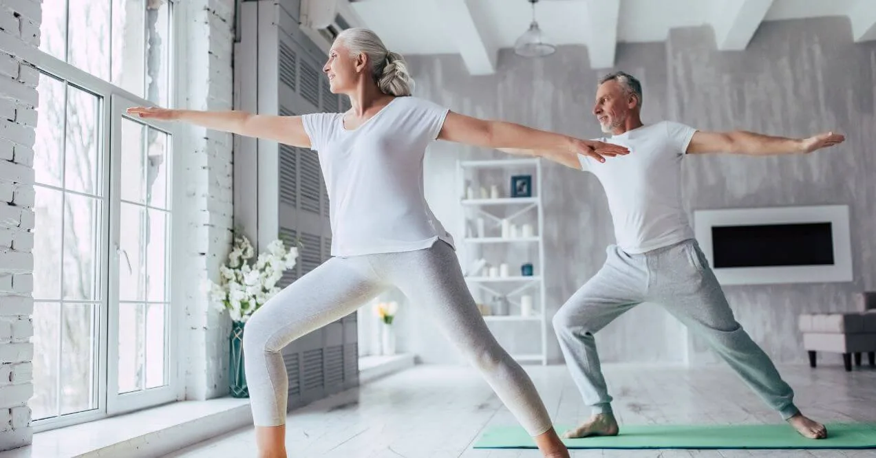 Senior couple practicing somatic yoga in a bright room, stretching and balancing in a yoga pose near a large window.