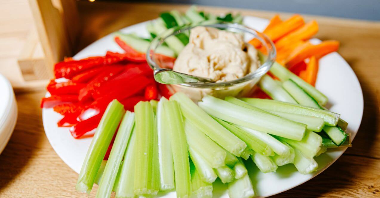 A plate of fresh celery, carrots, and red peppers with hummus dip.