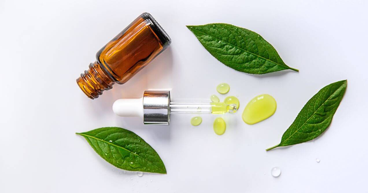 Tea tree oil bottle with dropper surrounded by green leaves and oil droplets.