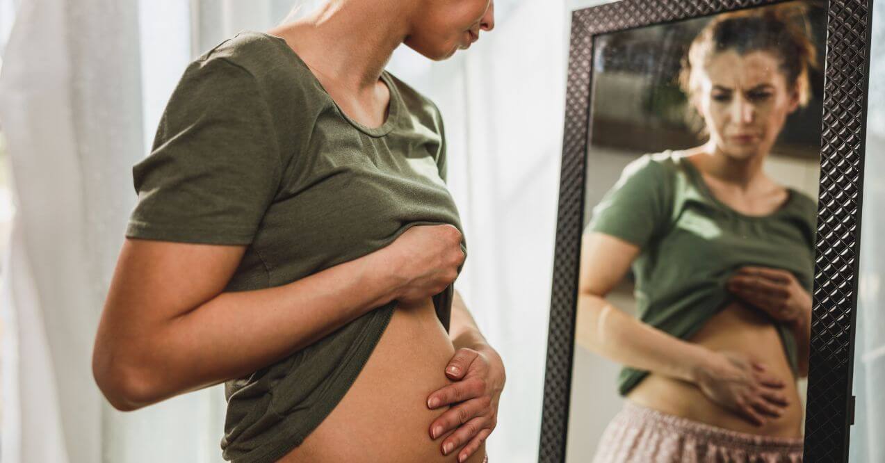 Woman examining her bloated stomach in front of a mirror.