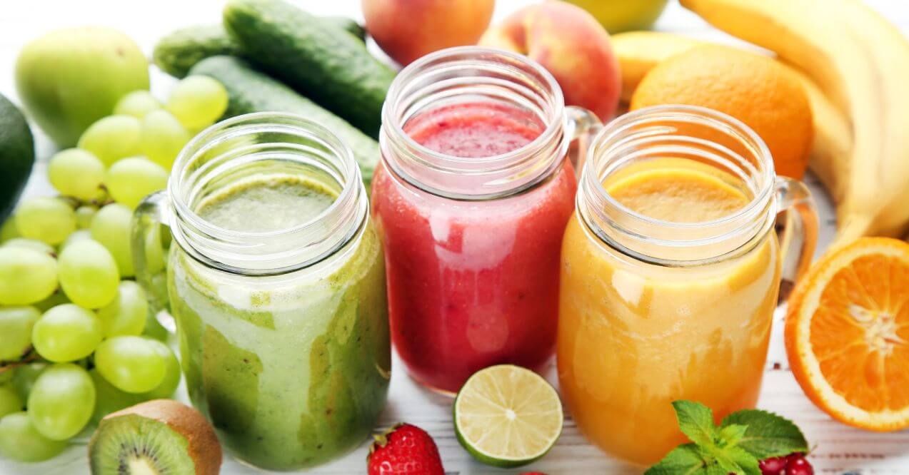 Three colorful fresh juice jars with fruits and vegetables in the background.