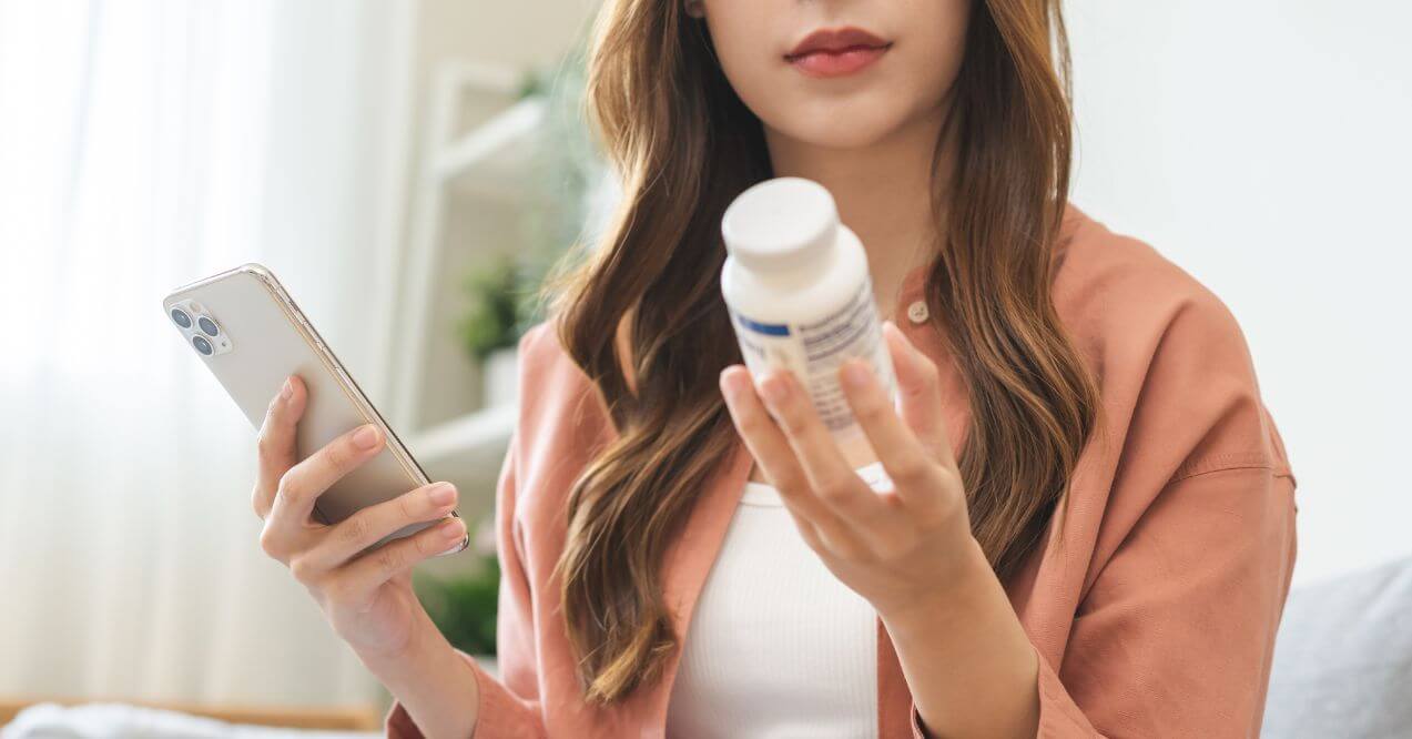 Woman reading a supplement bottle while researching on her phone.