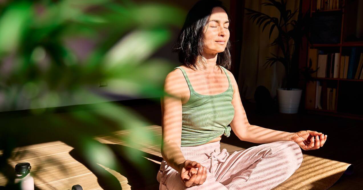 Woman meditating in a sunlit room with plants and soft shadows.