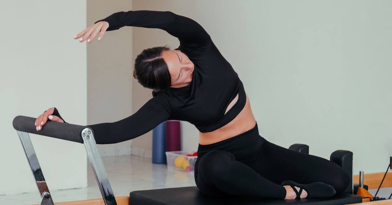 Woman practicing pilates, stretching on a reformer machine in a workout room.