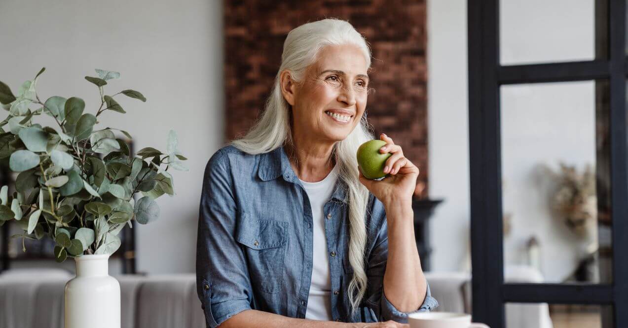 Smiling older woman holding a green apple while sitting at a table.