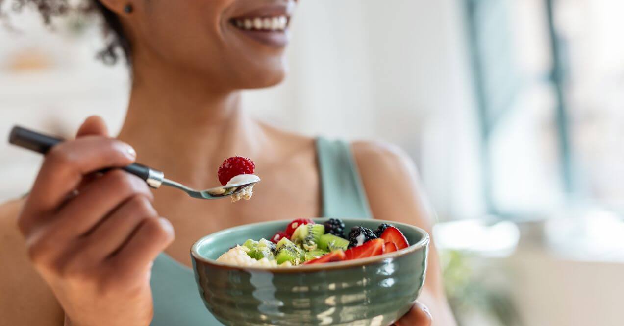 Person enjoying a bowl of yogurt topped with fruits like strawberries and kiwi.