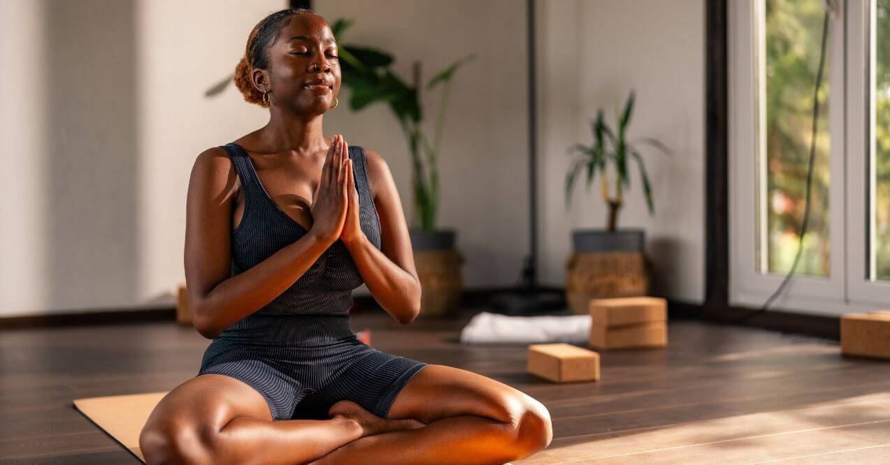 Woman meditating on a yoga mat in a sunlit room with plants and yoga blocks.