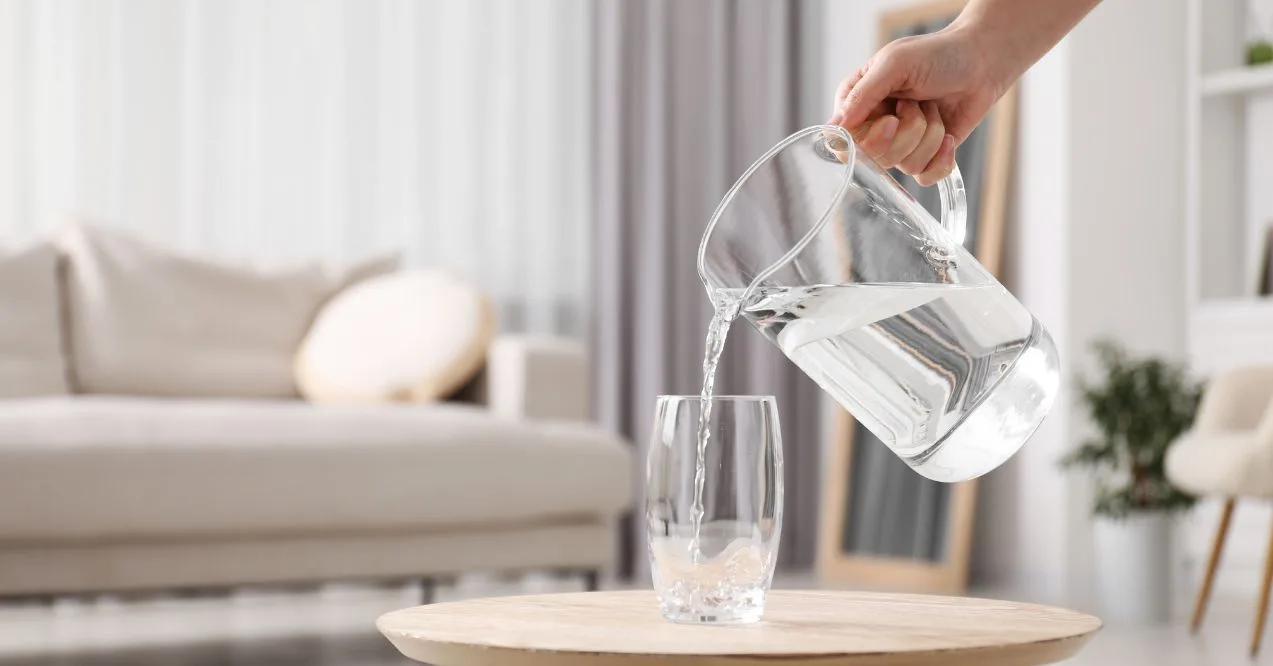 Hand pouring water from a pitcher into a glass on a table.