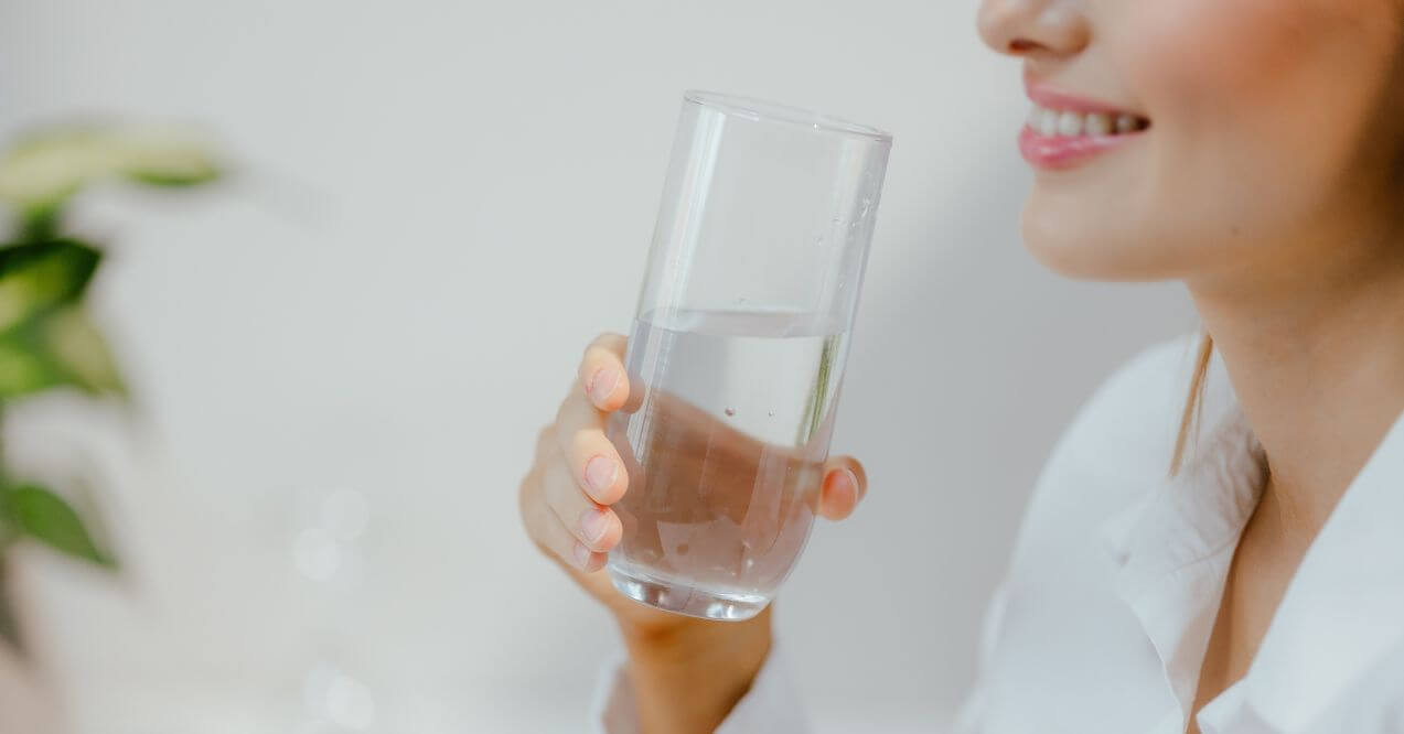 Close-up of a woman smiling while drinking a glass of water to stay hydrated.