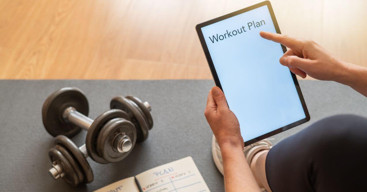 Person holding a tablet with "Workout Plan" on the screen near dumbbells and a notebook.