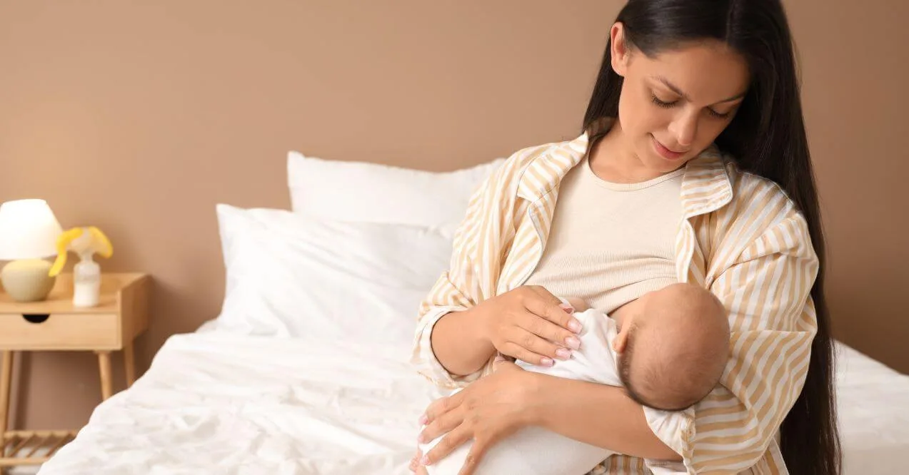 Mother in a striped robe breastfeeding her baby while sitting on a bed.