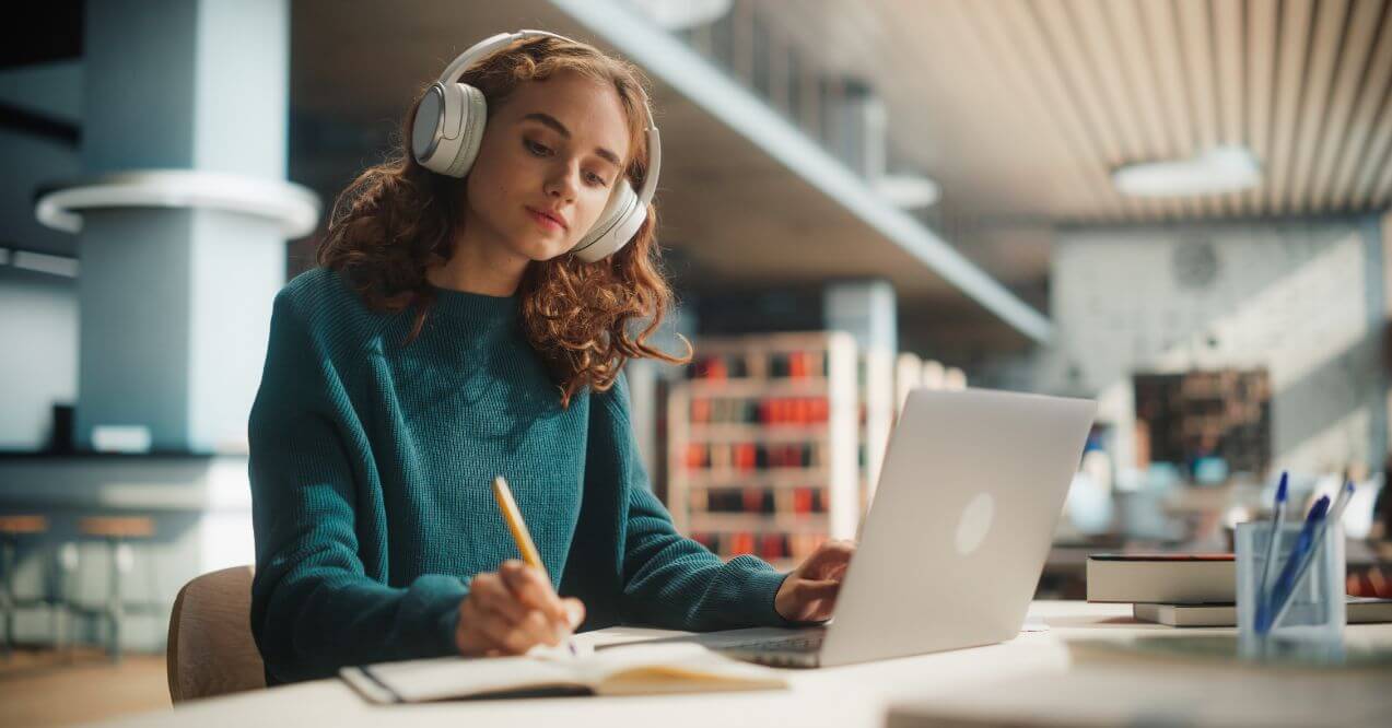 Woman studying in a library with headphones on, using a laptop and notebook.