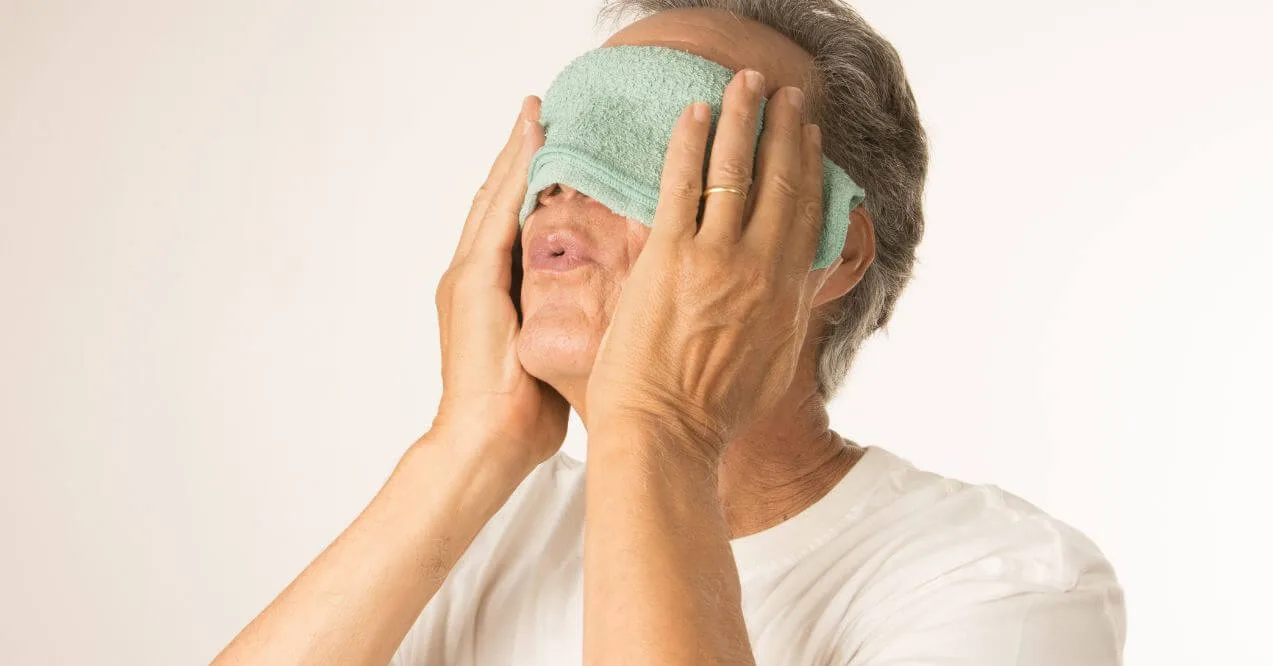 Elderly man applying a warm towel compress to his eyes.