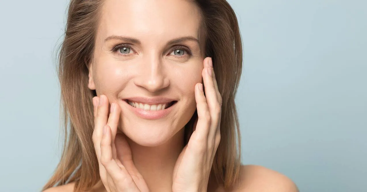 Smiling woman touching her glowing skin with both hands against a light blue background.