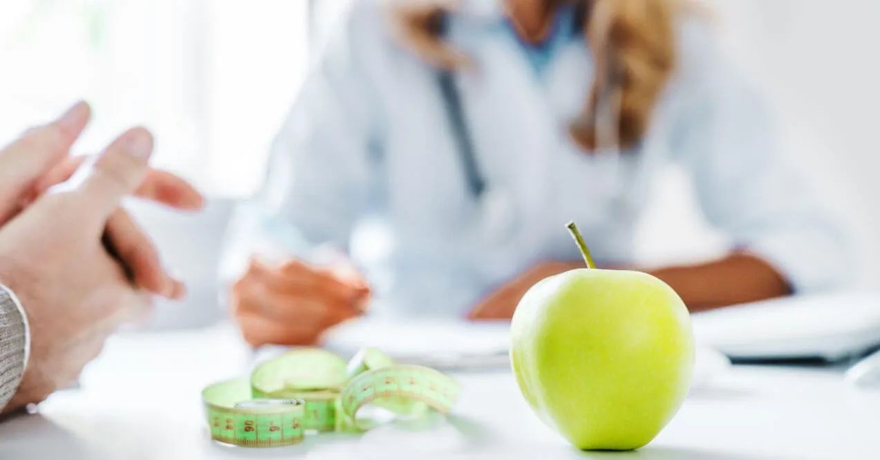 Green apple with a measuring tape on a table, with a blurred doctor in the background.