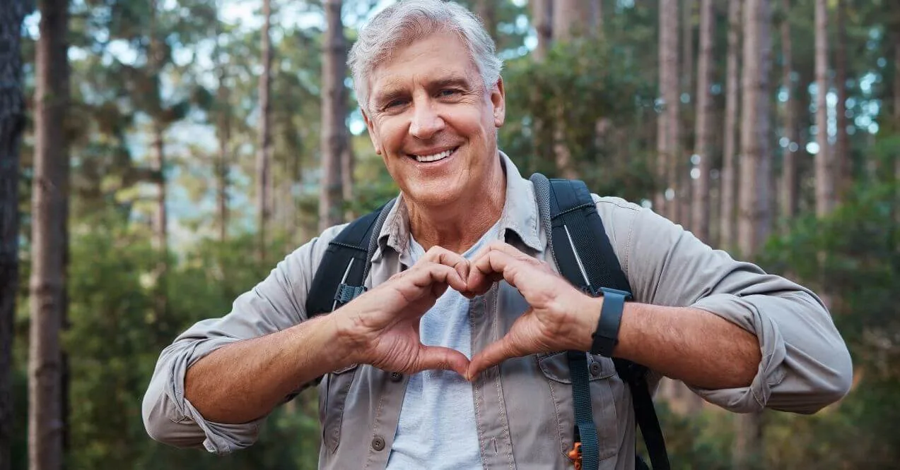 Older man in a forest making a heart shape with his hands and smiling.