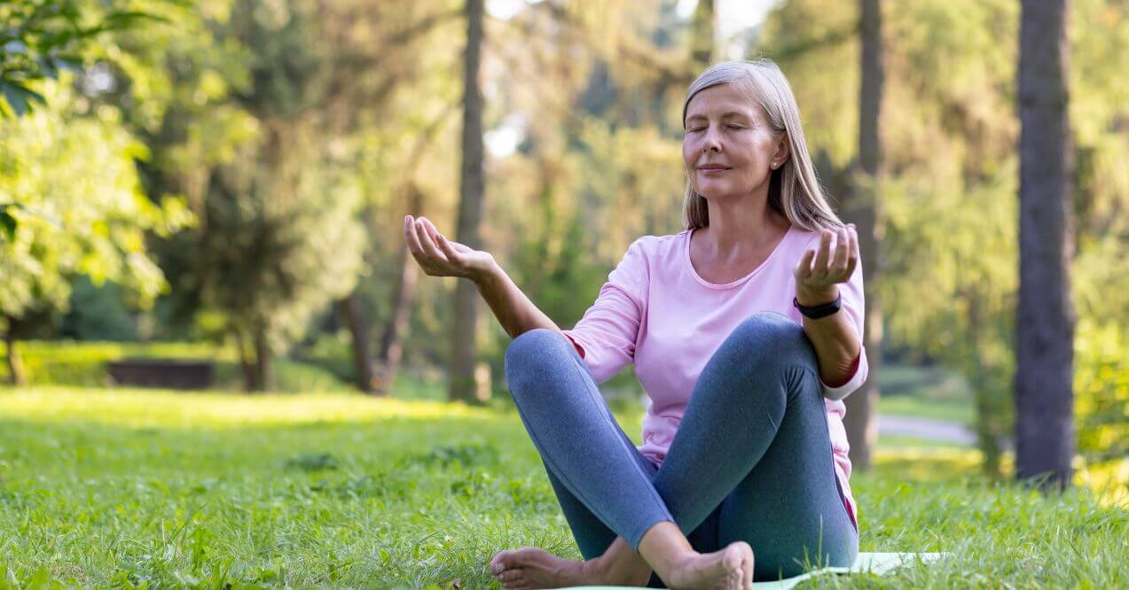 Older woman meditating outdoors on grass, surrounded by trees and natural light.