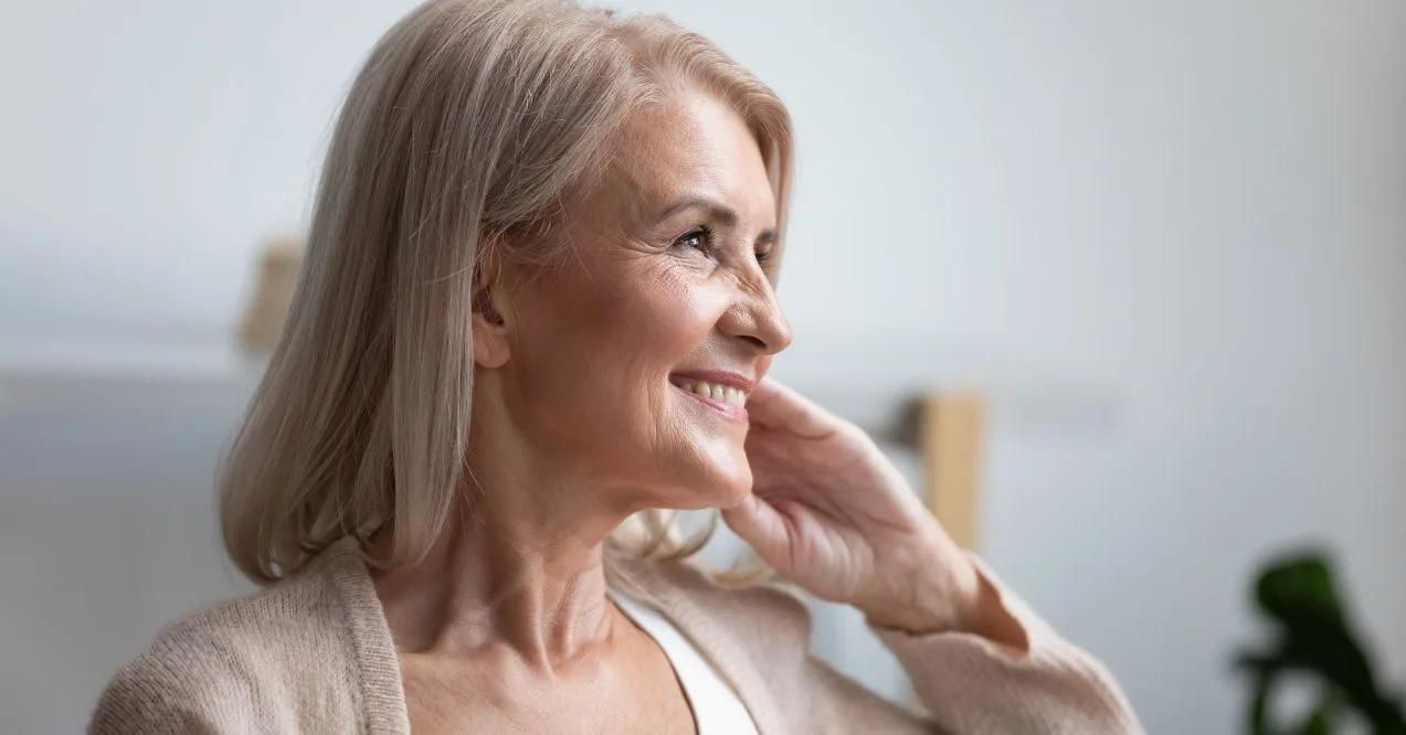 A mature woman with silver hair smiling while touching her neck in a relaxed manner.