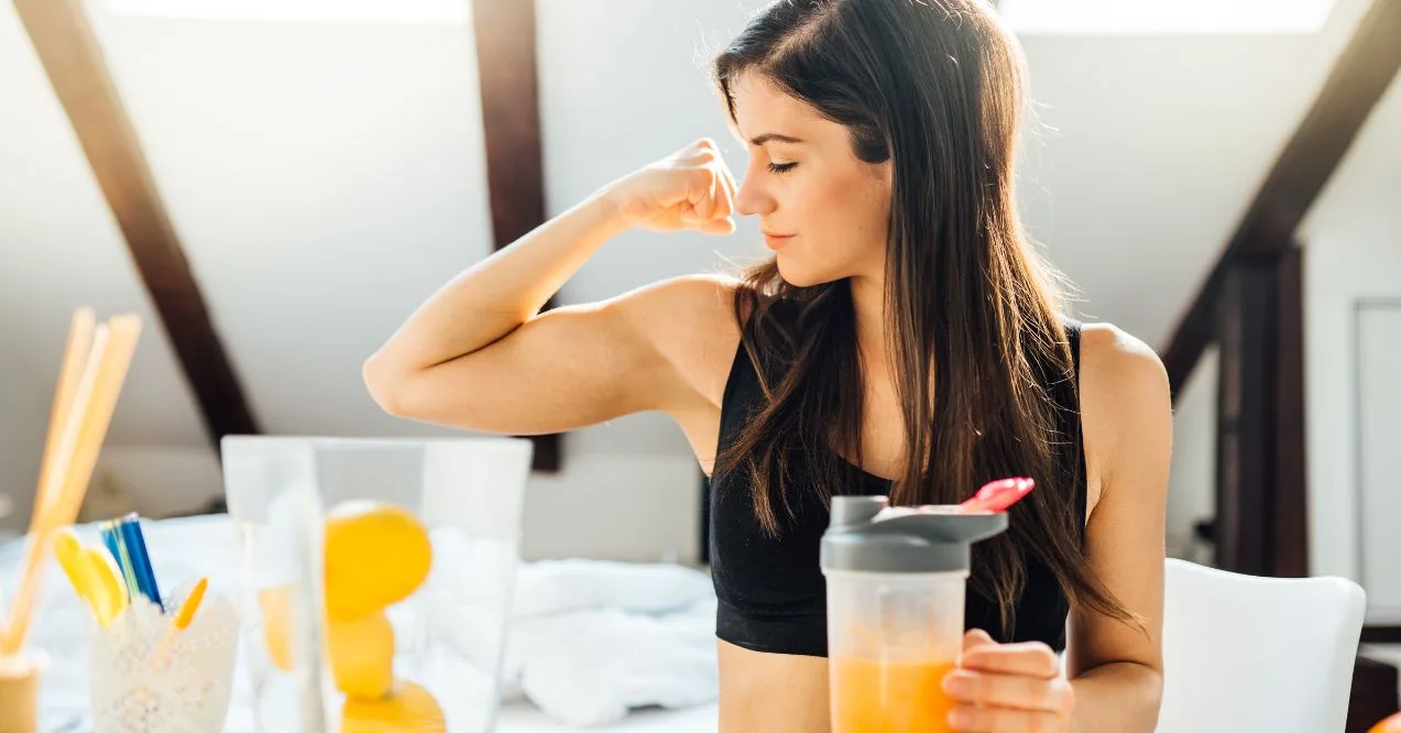 A fit young woman flexing her bicep while holding a glass of orange juice in a bright room.