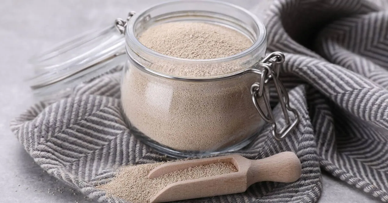Glass jar filled with yeast on a gray cloth with a wooden scoop beside it