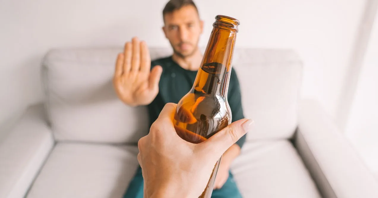 A man sitting on a couch raises his hand, refusing a bottle of beer.