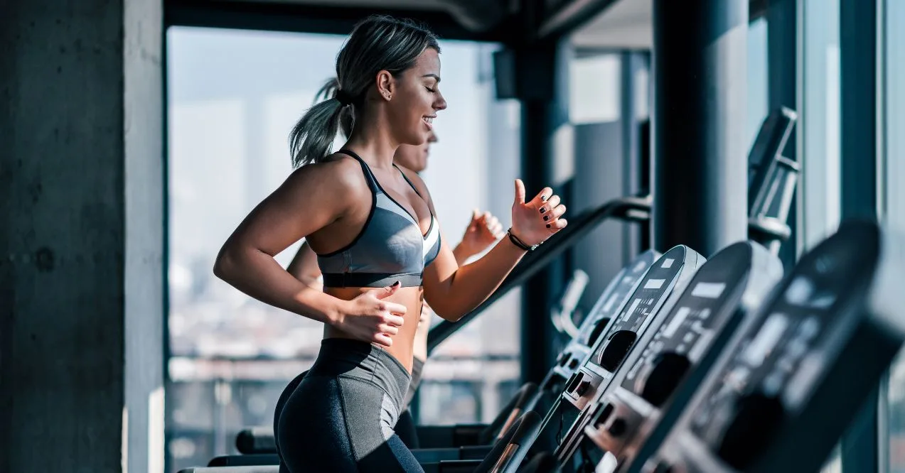 A woman running on a treadmill in a modern gym with large windows and natural light.