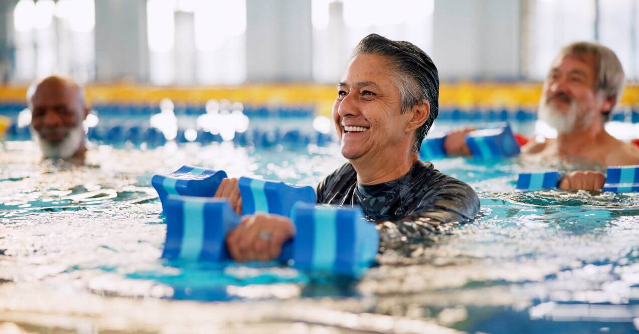 Older adults participating in water aerobics with foam weights, smiling and enjoying exercise and wondering does swimming help aging.