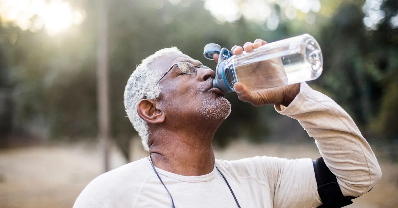 A senior man outdoors drinking water from a bottle after exercise, wearing a workout outfit.
