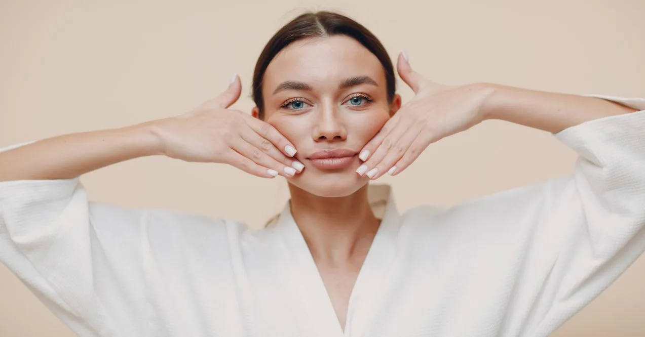 A woman in a white robe performing facial exercises with her hands on her cheeks.
