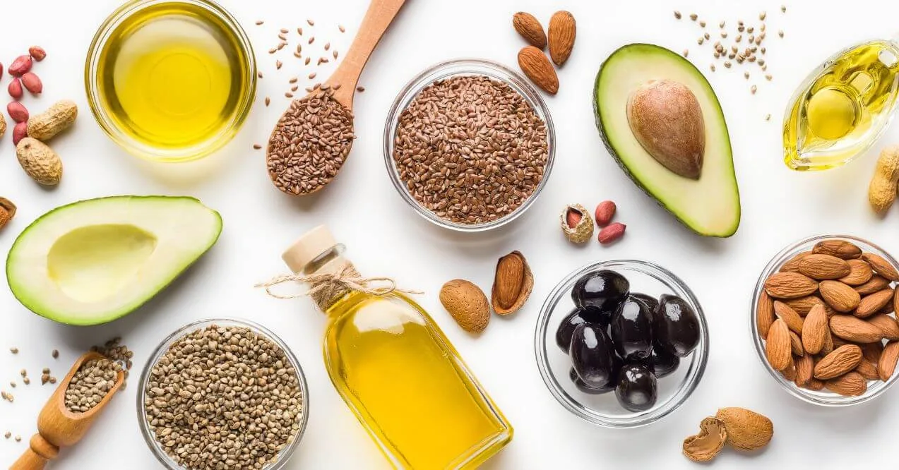 Various healthy fats, including avocados, nuts, seeds, and oils, displayed on a white surface.
