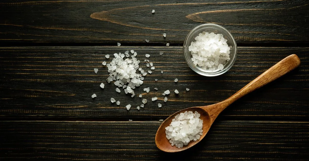 Coarse salt scattered on a dark wooden surface with a wooden spoon and a small glass bowl.