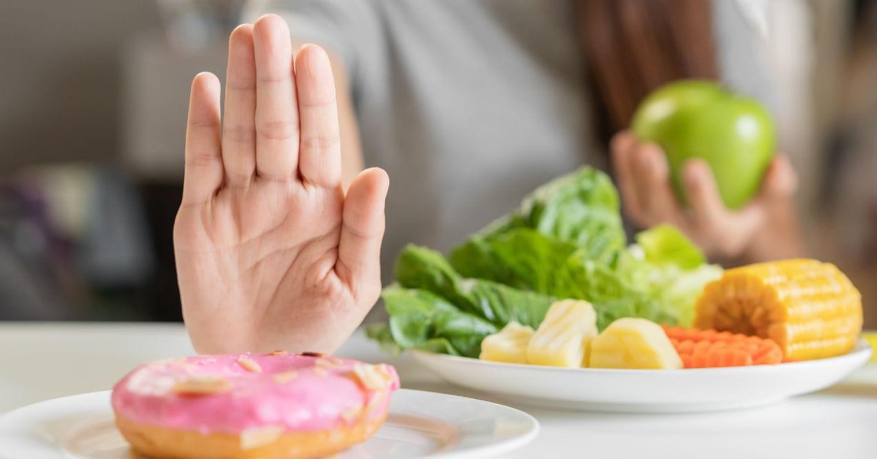 Hand rejecting a pink donut while choosing healthy foods like vegetables, fruits, and corn resembling what foods to avoid if alkaline phosphatase is high.