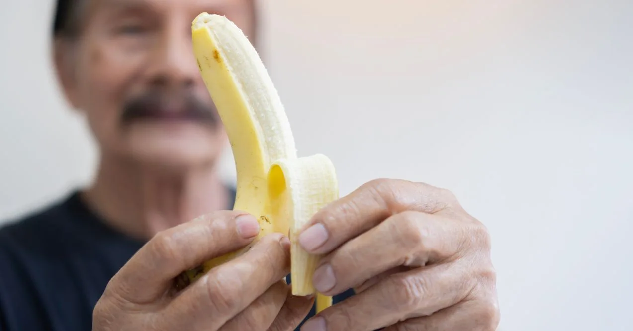 An elderly man peeling and holding a ripe banana close to his face resembling a question does eating bananas help with lymphedema